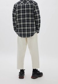 Persona in piedi di spalle, che indossa una camicia a quadri bianca e nera, pantaloni larghi color avorio, calzini neri e scarpe da ginnastica nere con suola rossa.