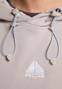 Hoodie gris clair avec cordons de serrage, présentant des accents bleus. Affiche un logo ACG brodé. Tissu doux et texturé avec un design décontracté.