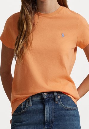 Femme portant un t-shirt orange à manches courtes avec un petit logo brodé et un jean bleu avec des poches visibles.