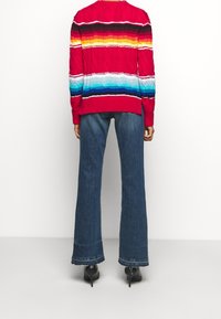 Pull en tricot rouge à rayures horizontales multicolores, associé à un jean denim bleu flare. Des talons noirs complètent la tenue.