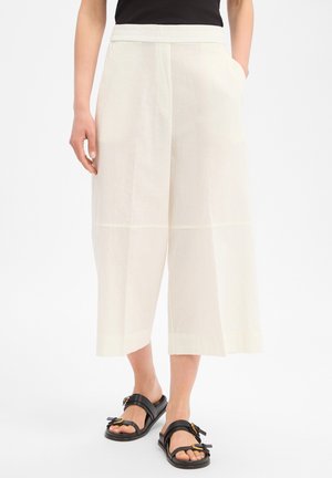 Vrouw draagt crèmekleurige, brede broek met hoge taille, die net boven de enkels is ingekort, en zwarte sandalen met open teen en gouden gespen, handen in de zakken.