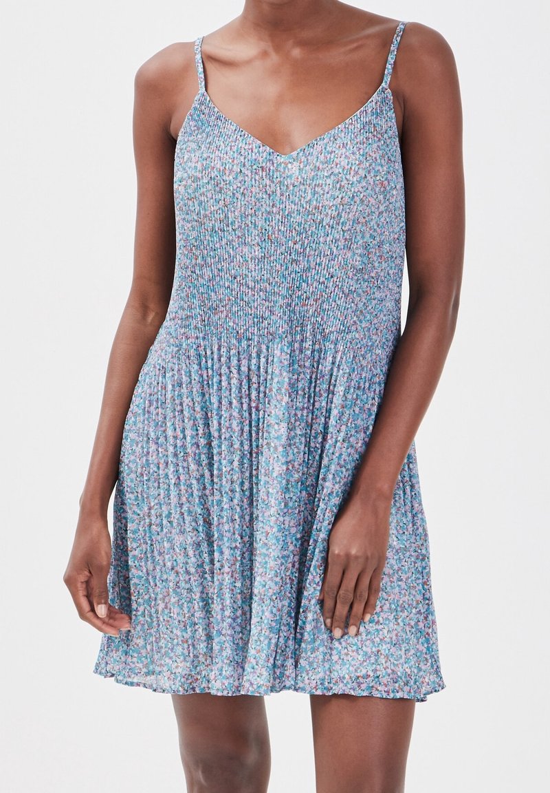 Femme portant une robe d'été bleue fleurie sans manches avec une jupe plissée et des fines bretelles.