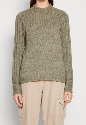 Femme portant un pull en maille côtelée vert olive avec des manches ballon et un pantalon cargo beige avec des poches latérales, debout devant un fond uni.