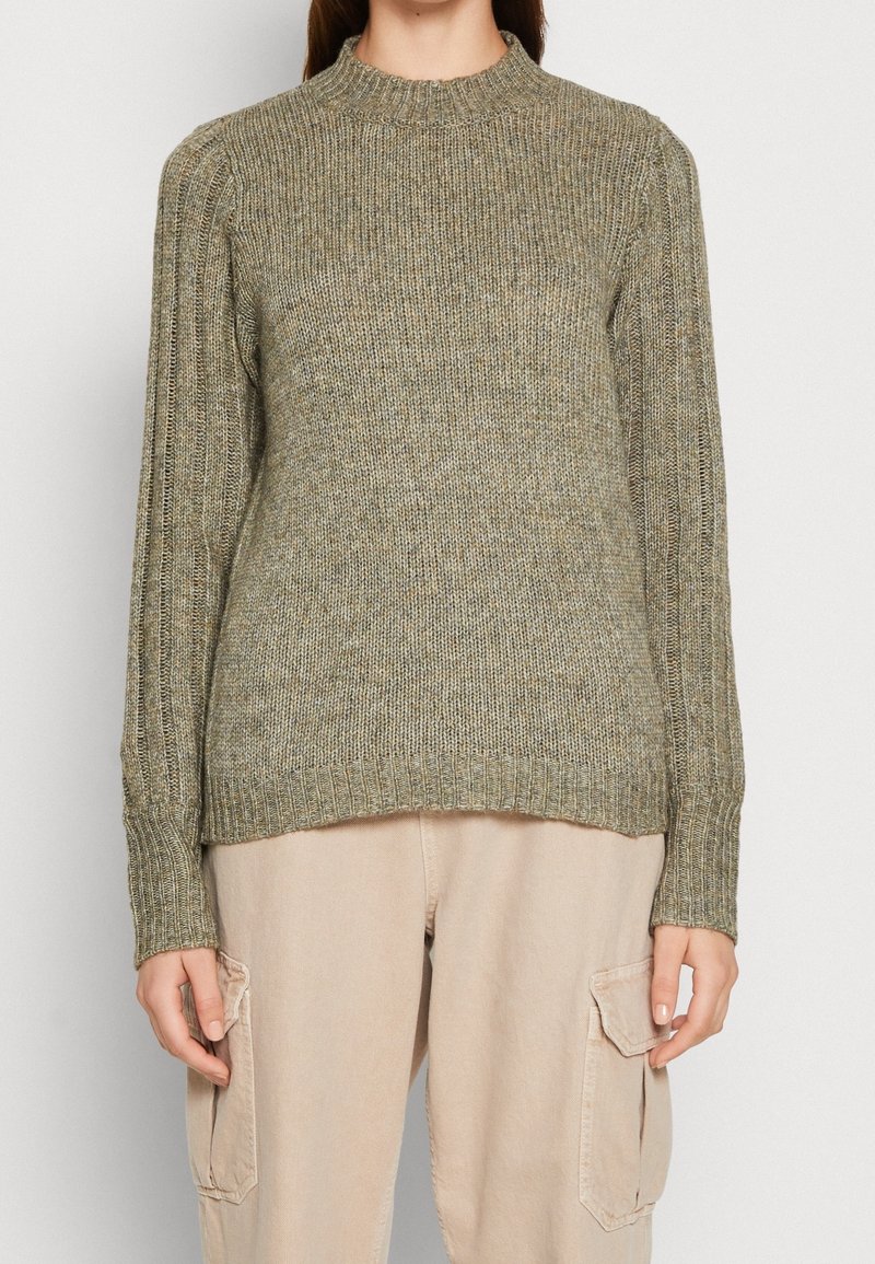 Femme portant un pull en maille côtelée vert olive avec des manches ballon et un pantalon cargo beige avec des poches latérales, debout devant un fond uni.