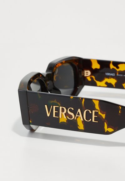 Black and amber tortoiseshell sunglasses with a chunky design, featuring gold "VERSACE" logo and bold patterns on the side temples.