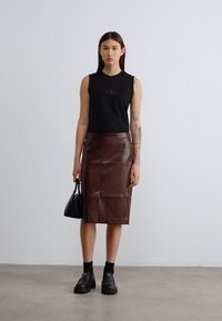 Μαύρο αμάνικο τοπ με λογότυπο, καφέ δερμάτινη φούστα μήκους μέχρι το γόνατο, ψηλά μαύρα παπούτσια, κρατώντας μια μικρή μαύρη τσάντα. Απλό φόντο.