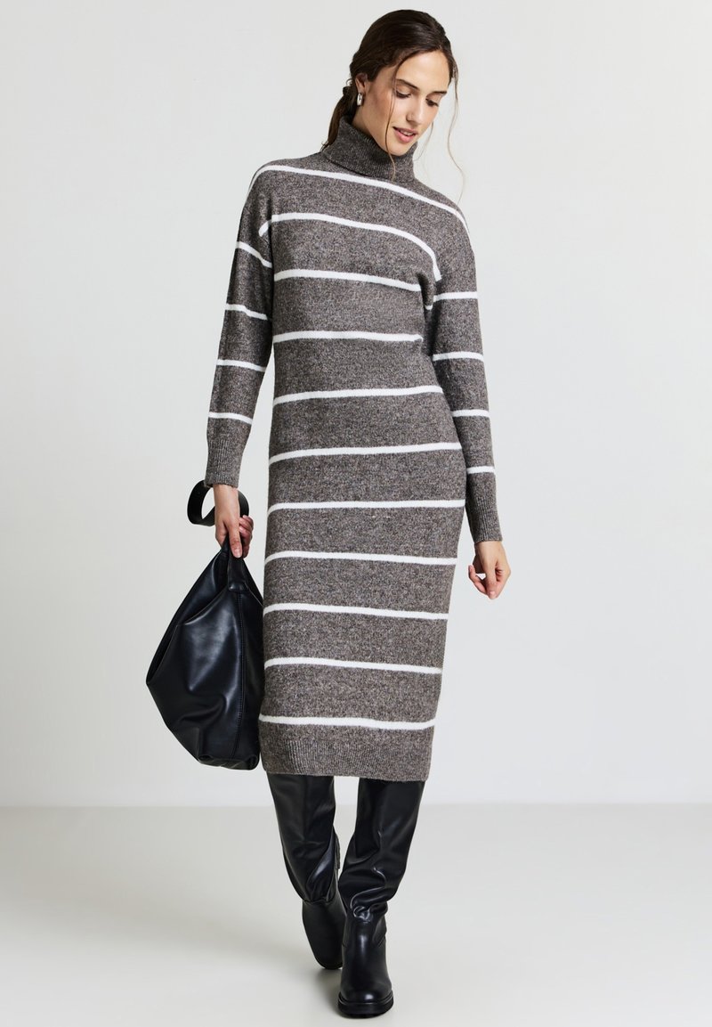 Abito grigio a maglia con strisce orizzontali bianche, colletto alto, maniche lunghe e design aderente, abbinato a una borsa tote nera e stivali neri.