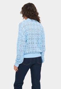 Gaiši zils adīts džemperis ar atvērto darbu rakstu, ribotām piedurknēm un apakšmalu, kā arī brīvā piegriezumā, apvienots ar tumšām džinsu biksēm.