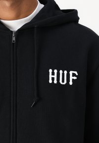 Hanorac negru cu fermoar, dintr-un material moale, având logo-ul brodat alb „HUF” pe piept, cu detalii de șnur și fermoar.