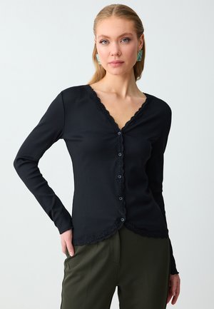Femme aux longs cheveux blonds porte un pull noir boutonné avec bordure en dentelle et un pantalon vert, debout devant un fond blanc uni.