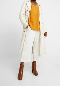 Trench coat beige con bottoni dorati, maglione giallo senape texturizzato, pantaloni bianchi a gamba larga e stivali alla caviglia marroni con tacco largo.