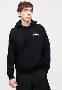 Μαύρο φούτερ με κουκούλα από βαμβάκι, με μπροστινή τσέπη καγκουρό, κορδόνια και λευκό λογότυπο "OBEY" στο στήθος. Άνετη εφαρμογή.