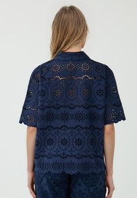 Una persona con capelli lunghi indossa un top in pizzo blu navy con intricati motivi ritagliati, visto da dietro su uno sfondo chiaro.