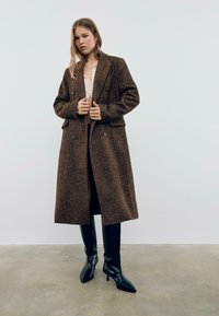 Manteau en chevrons brun avec des boutons double boutonnage et des poches à rabat, associé à des bottes noires jusqu'au genou et un haut de couleur claire.