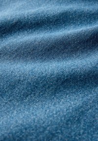 Denim w średnim odcieniu niebieskiego, charakteryzujący się tkanym splotem w skośną krateczkę, z widocznymi szwami, zagięciami i lekkim połyskiem.