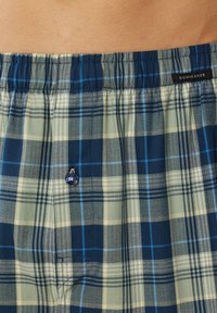 Boxers en tartan fabriqués en tissu léger, avec des couleurs bleu, vert et crème, une taille élastique et un petit bouton décoratif.