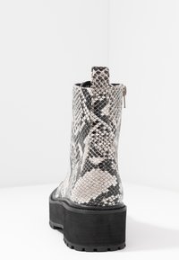 Botín hasta el tobillo con patrón de serpiente en blanco y negro, suela negra gruesa y un tirador de cremallera en la parte trasera sobre una superficie blanca.