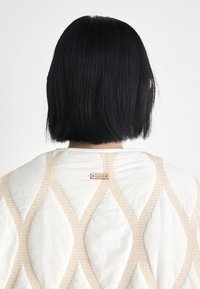 Quilted garment featuring a cream-colored fabric with beige diamond patterns and a metal tag reading "sesain" on the back.