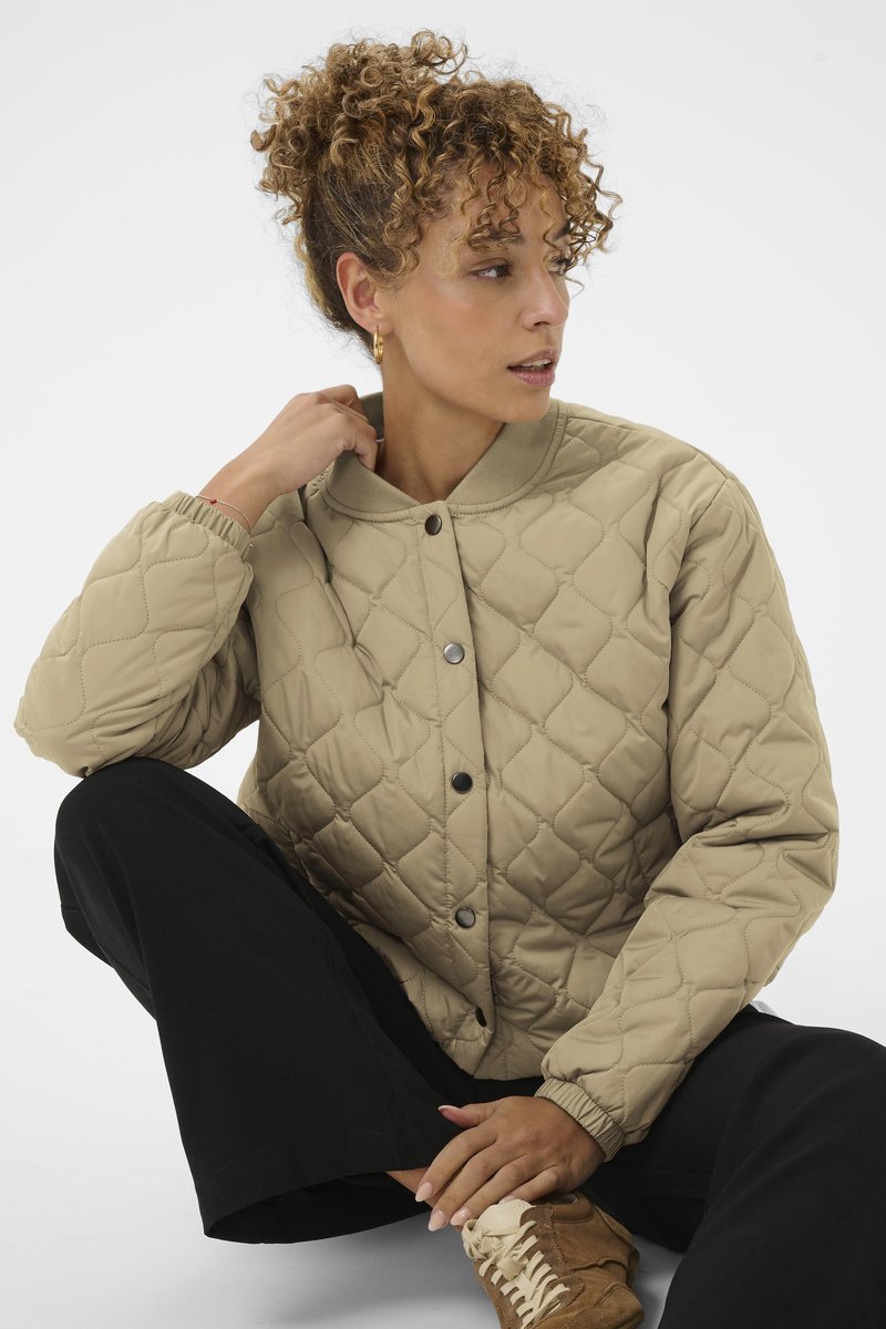 Beige gewatteerde jas met een ronde hals en knoopsluiting. Voorzien van elastische manchetten en een ruitpatroon, gecombineerd met zwarte broek en bruine schoenen.