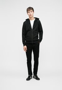 Schwarzer Kapuzenpullover mit Reißverschluss und zwei Fronttaschen, kombiniert mit schwarzen, schlank geschnittenen Hosen. Das Modell trägt schwarze Sneakers mit weißen Streifen. Darunter ein schlichtes weißes Hemd.