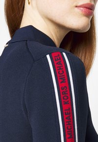 Gros plan d'une personne portant un haut bleu marine avec un détail d'épaule rayé rouge et blanc affichant le texte "MICHAEL KORS".