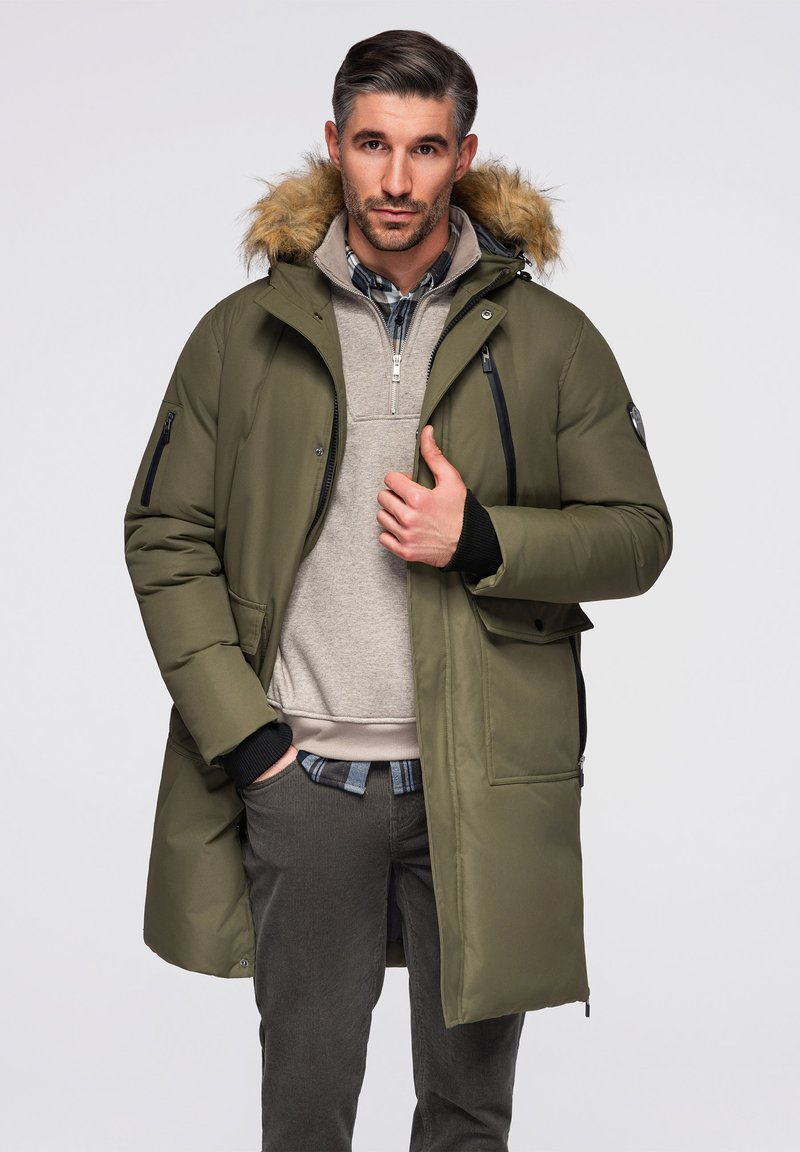 Mann trägt eine grüne Winterparka mit Fellkapuze, beigen Reißverschluss-Pullover, kariertes Hemd und graue Hose, steht mit einer Hand in der Tasche.
