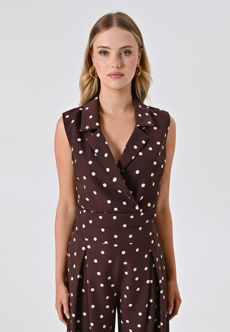 Jumpsuit senza maniche marrone con pois bianchi, dotato di colletto, scollo a V e dettagli plissettati in un tessuto morbido.