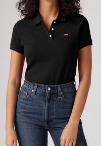 Černé polo tričko s límečkem a třemi knoflíky, na hrudi má malé červené logo Levi's, doplněné o džínsy s vysokým pasem v tmavém provedení.