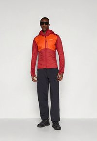 Homem vestindo casaco vermelho e laranja com capuz, calças pretas, sapatos pretos e óculos de sol escuros, posando contra um fundo cinza claro e simples.