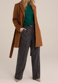 Bruine wollen jas, groene trui en grijze broek met wijdere pijpen en patroon. Beige puntige schoenen, riem met zilverkleurige details. Gestructureerde stoffen.