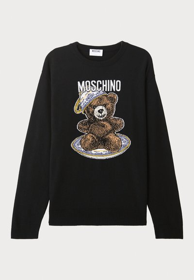 Melnā adīta džemperis ar lāča grafiku, kurš nēsā cepuri, apjozts ar šķīvja dizainu. Virs lāča ir uzraksts "MOSCHINO".