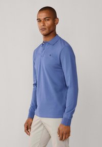 Dlhé rukávy polokošeľa v svetlomodrej bavlne, s golierovým výstrihom, zapínaním na tri gombíky a vyšívaným logom na hrudi.