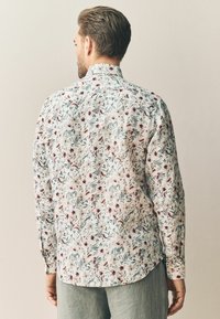 Chemise à manches longues en tissu léger, avec un motif floral dans des teintes de rose et de vert sur un fond blanc.