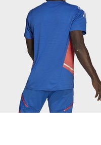 Modré sportovní tričko s červeným akcentem na boku, krátké rukávy a kulatý výstřih. Tkanina vypadá hladce a má mírně přiléhavý střih.