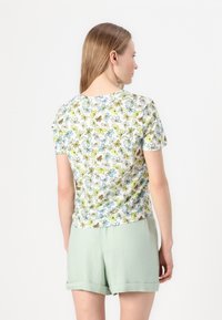 Kortermet hvit t-skjorte med blomsterdesign i grønn og blå, kombinert med lysegrønne shorts. Materialet er en blanding av bomull og gir en myk tekstur.