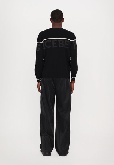 Pull en mailles noires avec des lettres grises dans le dos, accompagné d'un pantalon noir ample en matière brillante. Rayures blanches horizontales aux poignets.