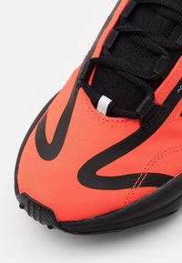 Jasně oranžová sportovní bota s elegantním designem, černými akcenty, texturovaným svrškem z látky a výrazným černým logem. Pryžová podrážka.