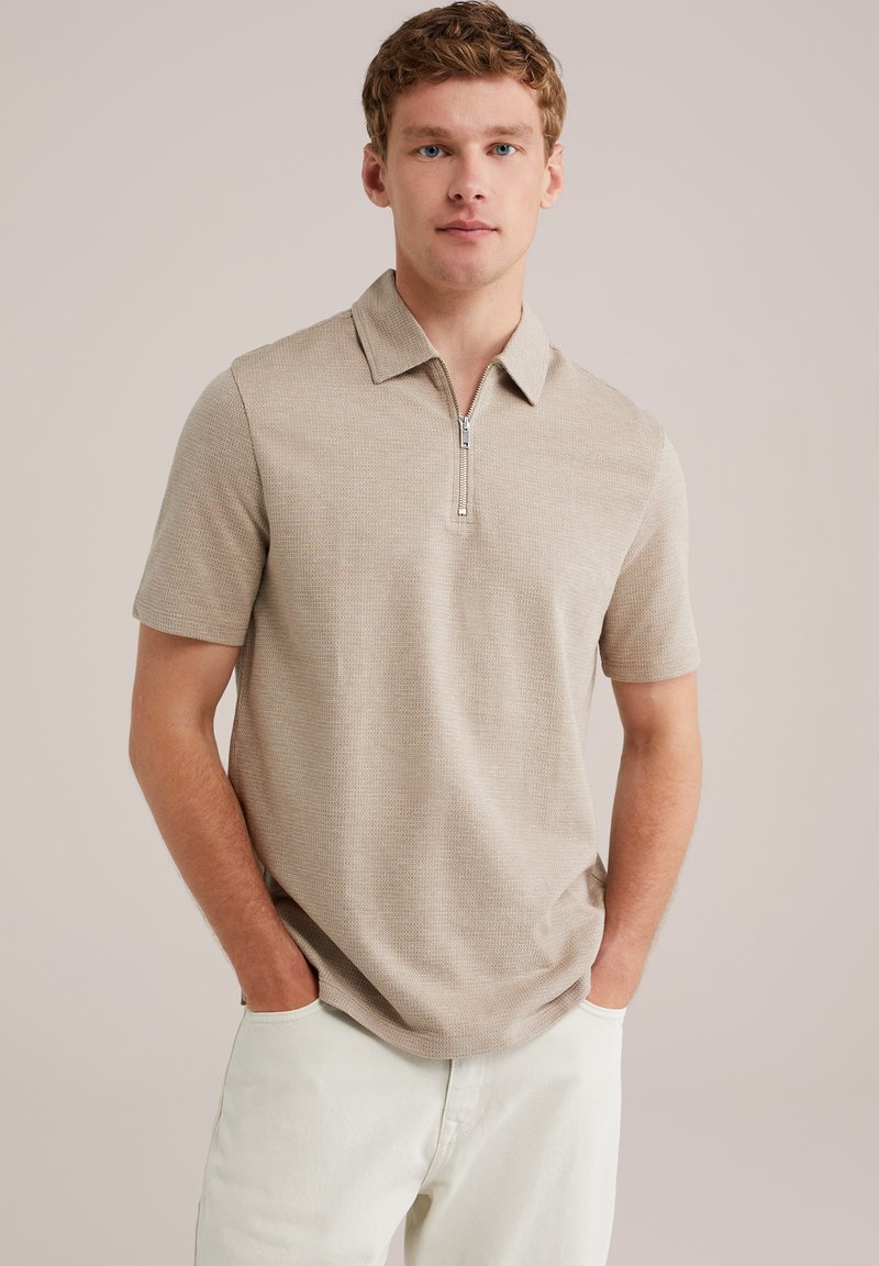 Camisa polo de manga corta beige con cuello con cremallera, con una tela texturizada y un corte relajado, combinada con pantalones de color claro.