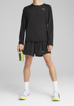 Persoon draagt een zwart top met lange mouwen en korte broek, houdt een gele Puma waterfles vast, draagt witte sokken en witte Puma sneakers.