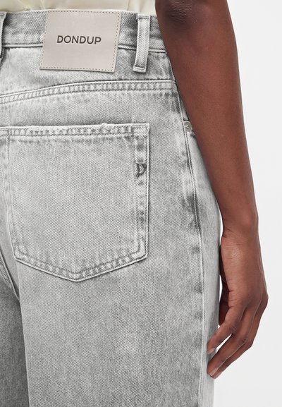 Gros plan de la poche arrière d'un jean en denim gris clair avec une étiquette de la marque Dondup et la main d'une personne à la peau foncée reposant à côté du jean.