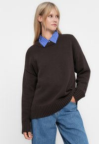 En blond kvinne iført en brun strikkegenser over en blå og hvit stripete skjorte med krage og løse blå jeans, med en hånd i lommen.
