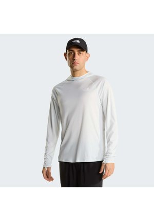 Hombre con sudadera con capucha blanca de manga larga y gorra negra, de pie con expresión neutral frente a un fondo claro y liso.