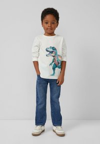 Weißes Langarmshirt mit einem bunten Dinosaurier-Graphic, kombiniert mit blauen Jeans und weißen Sneakers mit beigen Akzenten.