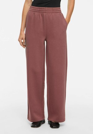 Femme portant un pantalon de jogging ample mauve taille haute avec ceinture élastique et chaussures noires, debout avec une main dans la poche.