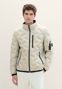 Helle beigen Steppjacke mit hohem Kragen, frontalem Reißverschluss und zwei Seitentaschen. Mit einer Reißverschlusstasche auf der Brust und schwarzen Akzenten.