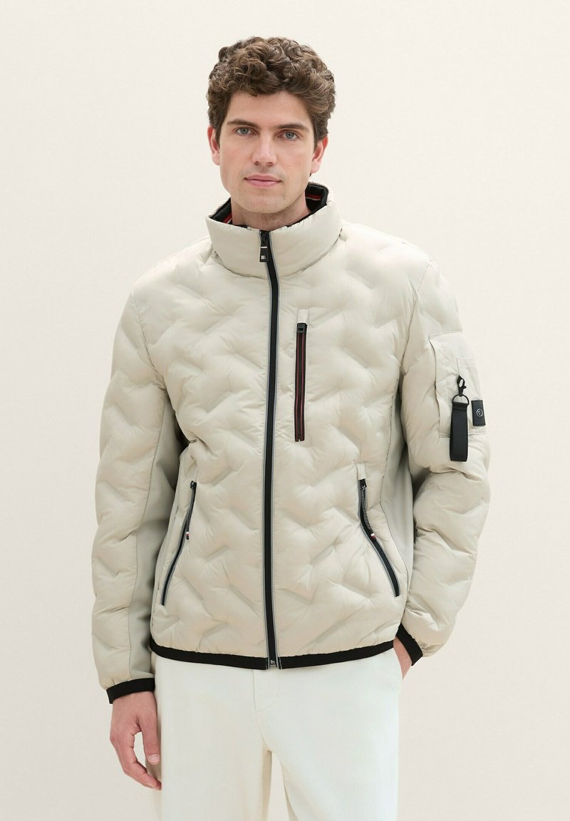 Helle beigen Steppjacke mit hohem Kragen, frontalem Reißverschluss und zwei Seitentaschen. Mit einer Reißverschlusstasche auf der Brust und schwarzen Akzenten.