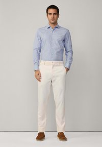 Camicia a righe blu chiaro con maniche lunghe, abbinata a pantaloni crema e scarpe in suede marrone. Bottoni semplici, vestibilità su misura.