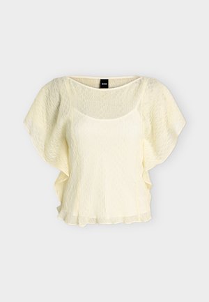 Blouse à manches courtes couleur crème avec délicate superposition de dentelle, col rond et ourlet à volants, conçue pour une coupe légère et aérienne.