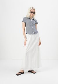 Kortermet, knappet skjorte i grått rutemønster kombinert med hvit, blondekjole og svarte flip-flops, modellen står foran en nøytral bakgrunn.