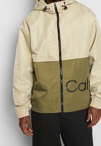 Lekka kurtka z kapturem o beżowej górnej części i oliwkowo-zielonej dolnej, z dużym nadrukiem "Cal", z przodu zapinana na zamek oraz z elastycznymi mankietami.